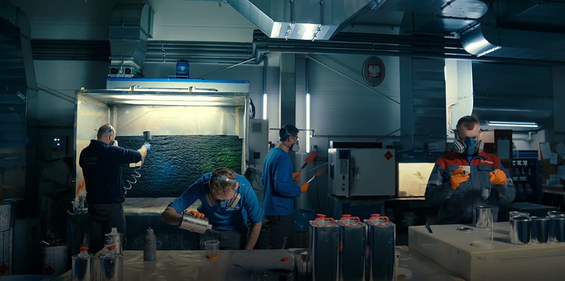 Workers in painting area performing preparatory work. Industrial room with lighting and ventilation system, four workers in blue work suits.