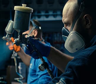 Close-up of painter in protective mask operating spray gun. Second worker in blue suit visible in background.