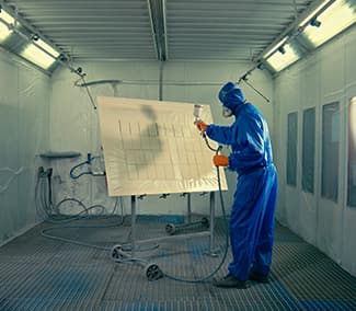 Worker in blue protective suit painting large white panel in professional paint booth equipped with ventilation and lighting system.