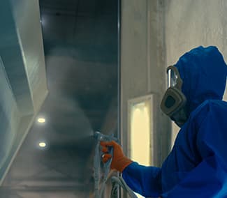 Worker in blue protective suit and gas mask performing spray painting in upper part of paint booth. Spot lighting visible in background.