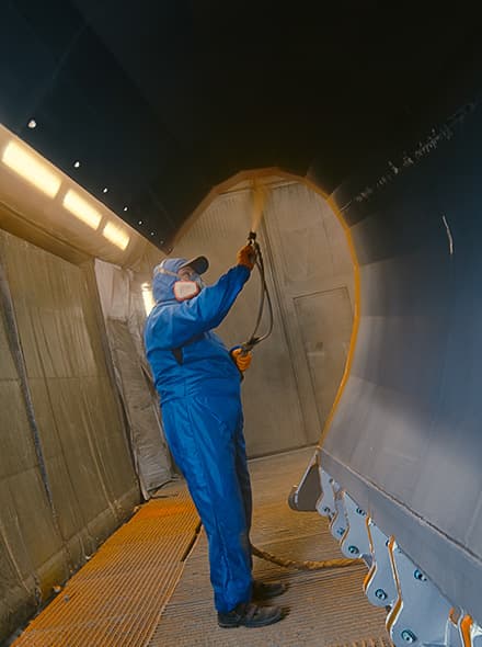 Worker in blue protective suit performing spray painting at height. Visible paint booth with ceiling lighting system.